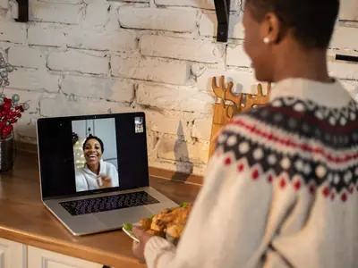 A person in a patterned sweater has a video call on a laptop in a kitchen.
