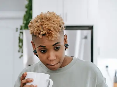 A Black woman with short curly hair holds a mug of coffee in her kitchen, lost in thought.