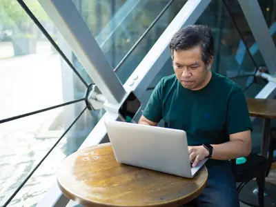 A middle-aged East Asian man types on a laptop at a round table in a glass-walled cafe.