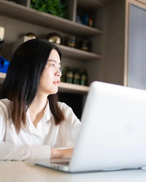 A young East Asian woman works on a laptop at a bright, tidy home office desk.