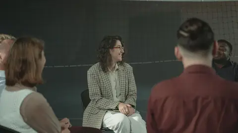 A small mixed group of Black and White adults sits in a circle of chairs, listening as one woman speaks.
