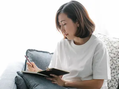 A woman sits on a gray couch writing in her diary, with a mug beside her.