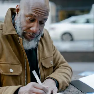 An older Black man with a grey beard writes in a notebook at an outdoor table.