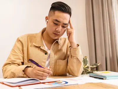 A young East Asian man writes notes at a desk, resting his head on his hand in thought.