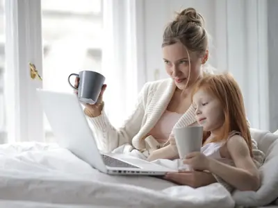 A mother with a coffee mug and a laptop shares a cozy morning in bed with her young daughter.