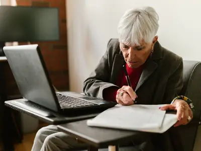 An older woman with short gray hair writes on paper beside an open laptop.
