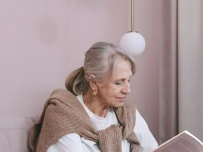 An older woman with gray hair and a cardigan over her shoulders reads a book on a couch.